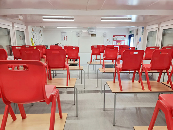 Modular classroom building interior with rows of desks and red plastic chairs, providing a temporary or permanent school classroom solution.