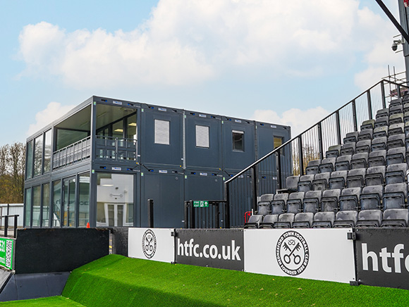 Modular sports facility building at a football ground, providing modern changing rooms, offices, and hospitality spaces alongside outdoor spectator seating.