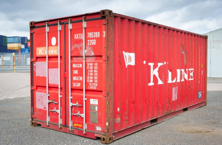 Weathered red steel shipping container with visible rust marks and white printed text including weight specifications and the brand name ‘K Line.’ The container has double doors with metal locking bars and is placed on an outdoor gravel surface near an industrial area with fencing and stacked containers in the background under a cloudy sky. 