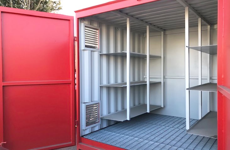 Interior of a converted shipping container fitted with long industrial storage shelves on the left and a countertop with sink and wall-mounted shelving on the right, brightly lit with white clean walls and windows. 