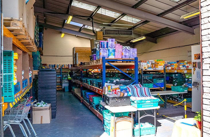 Interior of a food bank warehouse with stocked shelves of packaged goods and supplies, including crates and trolleys. 