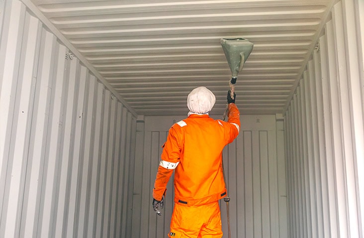 A person wearing an orange protective suit and a white head covering is applying coating to the ceiling of an empty shipping container using a spray tool. 