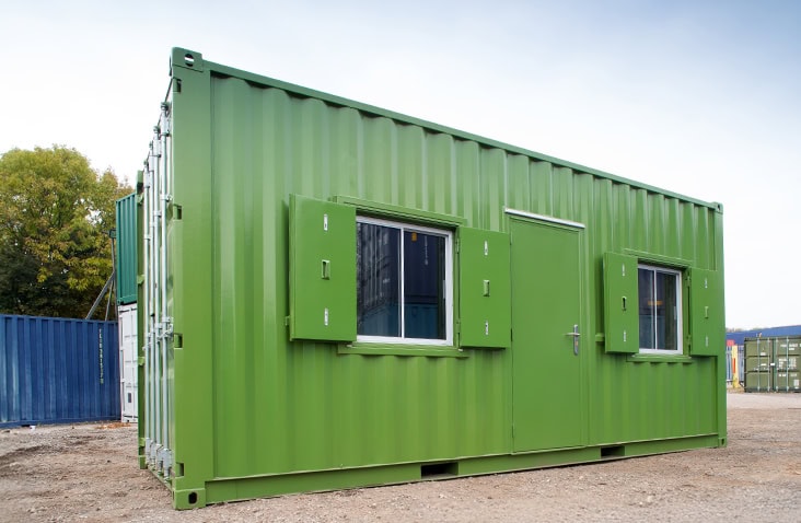 Green converted shipping container office with personnel door and two secure shuttered windows, positioned outdoors on a storage depot yard. 