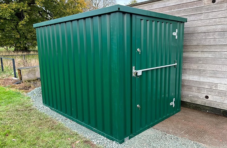     Green flat pack steel storage unit with single door and external locking bar, positioned outdoors beside a wooden building. 