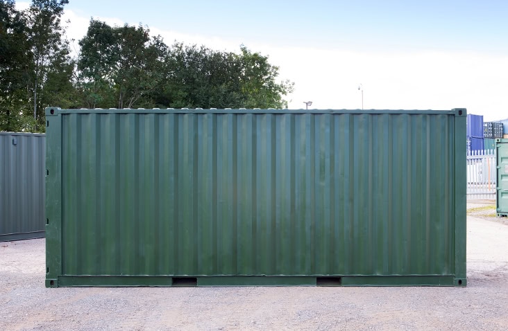 Refurbished 20ft shipping container painted dark green, shown from the side on a gravel yard with trees and other containers in the background.