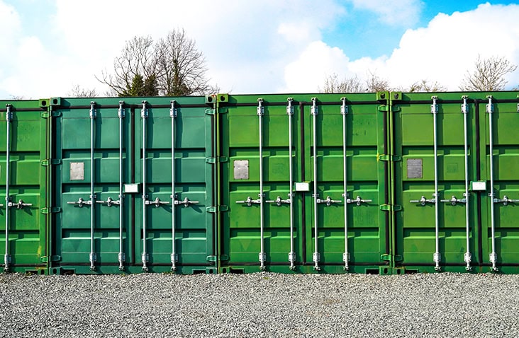 Row of green 20ft self storage containers for hire on a gravel surface, used for container storage solutions, with trees and sky in the background. 