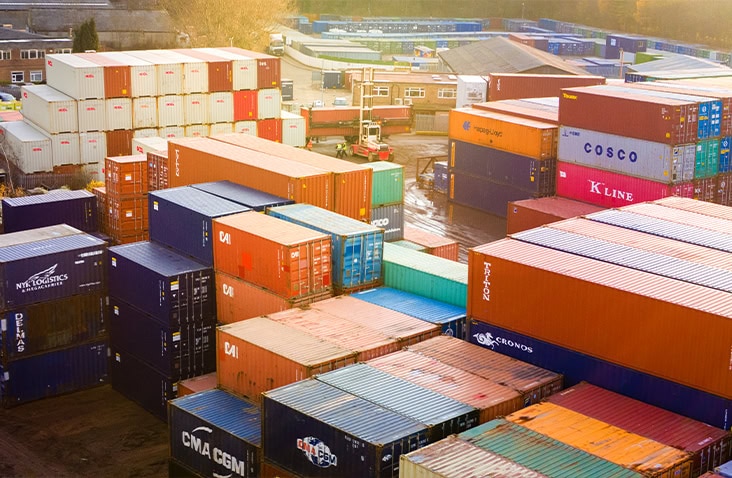 Drone photograph of a large shipping container depot filled with stacked multicoloured containers. A red container handler forklift is visible moving containers between tall stacks. The sky is bright and partly cloudy. 