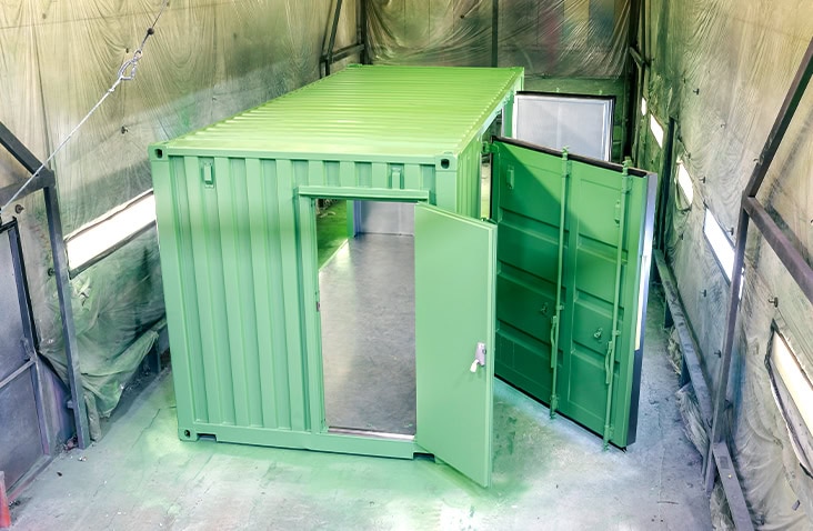 Freshly spray-painted green shipping container inside a paint booth, with both the main door and double cargo doors open, showing a clean interior. The booth walls are covered with protective sheets, and the floor has green overspray. 