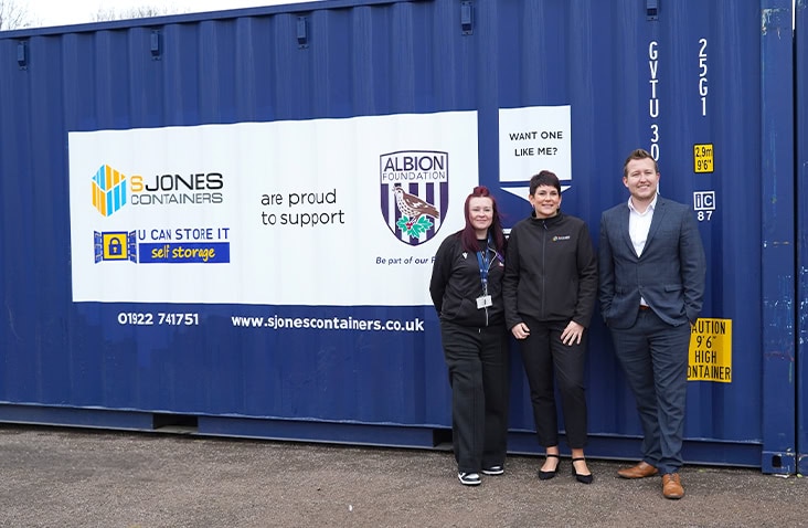     Three people standing in front of a blue 20ft shipping container branded with S Jones Containers, U Can Store It, and Albion Foundation logos, showing partnership support messaging. 