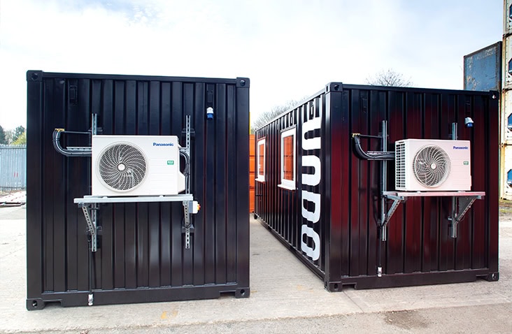 Two black converted shipping containers with externally mounted HVAC condenser units installed on steel brackets. 
