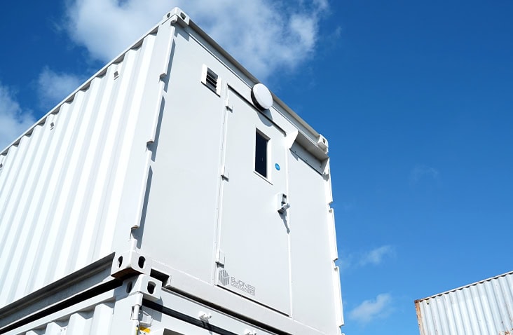 White converted shipping container with a personnel door and ventilation, stacked on another container against a blue sky. 