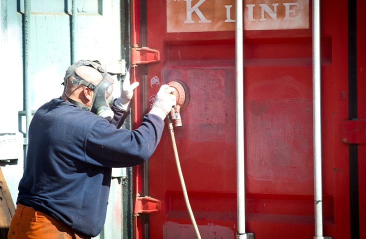 A person wearing protective gear and a respirator using a power tool to sand or grind the surface of a red shipping container door. The container has visible vertical locking bars and faded text reading ‘K LINE’ at the top. Bright sunlight illuminates the scene, with parts of other containers visible in the background.