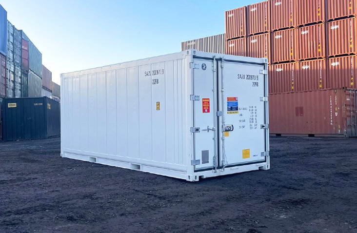 20ft white refrigerated shipping container shown from the front and side, with closed double doors displaying specification labels and safety signage, positioned outdoors in a container yard with stacked shipping containers in the background. 