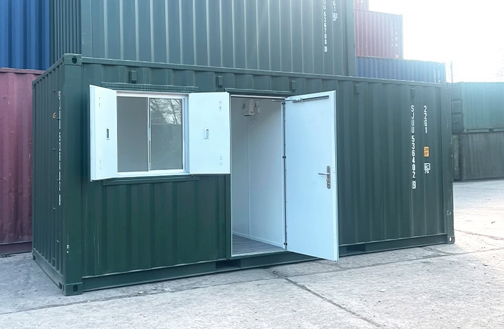 Converted green shipping container with open white personnel door and double window serving hatch, configured as an office and storage split. 
