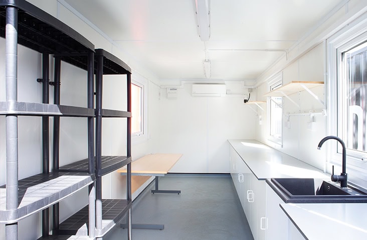 Interior of a converted shipping container with plastic shelving, workbench tables, white cabinetry, sinks, and windows, configured as a clean working lab space. 