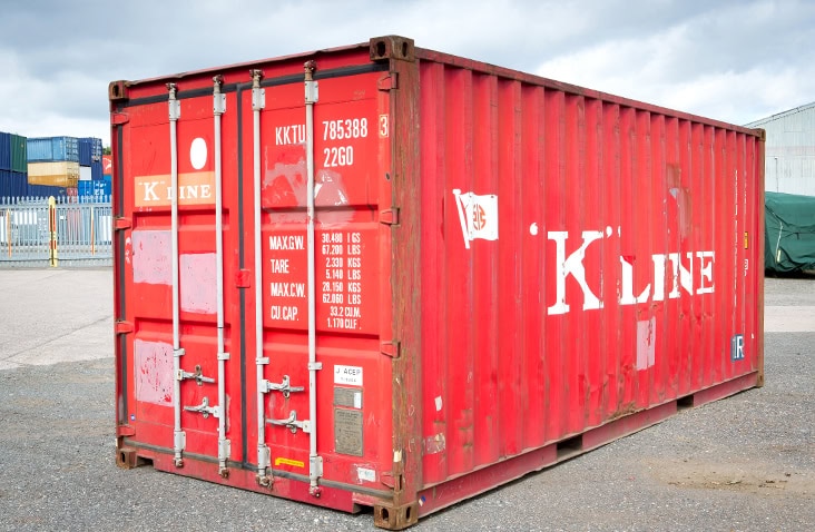     Used 20ft red shipping container with closed cargo doors, visible scuffs, rust patches, and faded branding, positioned on a yard surface with other containers in the background. 