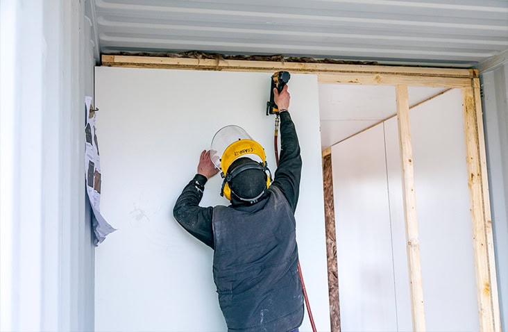 Technician installing an insulated internal partition wall inside a shipping container using a power tool and wooden framing. 
