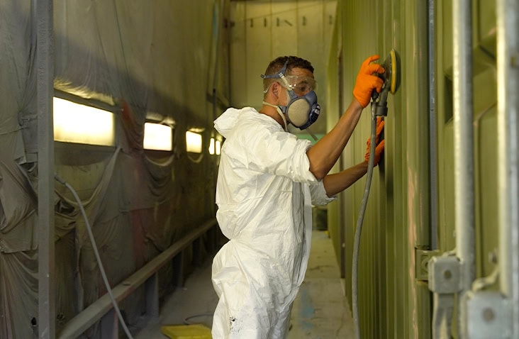 A person wearing a white protective suit, orange gloves, and a respirator is sanding the surface of a green shipping container inside a paint booth. 