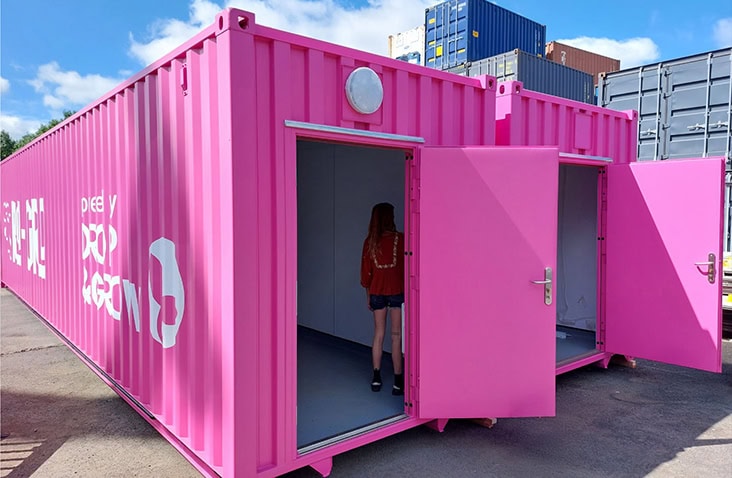 Bright pink converted shipping container with two open personnel doors, showing a person standing inside and window shutters open. 