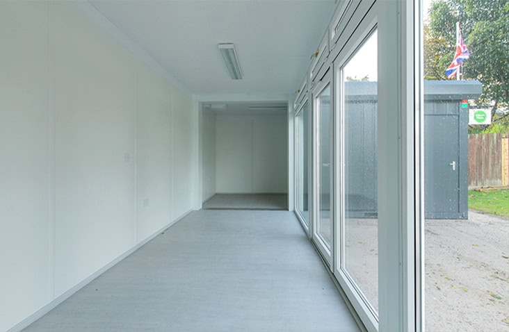 Interior of a modular building with white walls, grey flooring, ceiling lights, and large windows allowing natural light to fill the space.