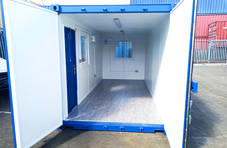 Interior of a blue converted shipping container office, viewed through open double doors. The space has insulated white walls, a personnel door, a window with a shutter, ceiling lighting, and a wood-effect vinyl floor.