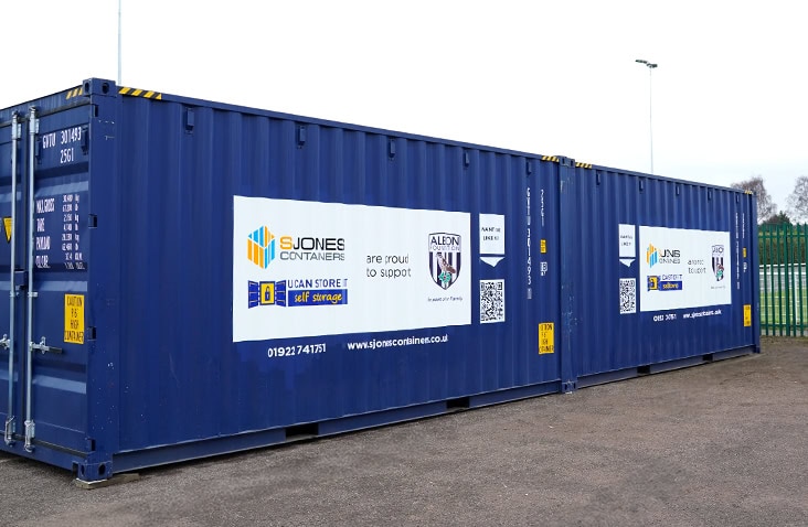     Two blue 20ft shipping containers positioned side by side on a sports facility site, featuring sponsorship branding for S Jones Containers and The Albion Foundation. 