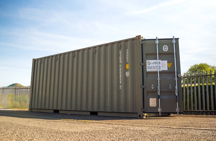 20ft green steel shipping container positioned on a yard beside metal fencing, branded with the S Jones Containers hire label. 