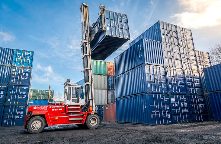 Large red container handler forklift lifting a green steel shipping container in a storage yard. The forklift raises the container high above stacked blue containers under a bright sky. 
