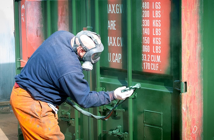 A person wearing protective gear and a respirator spray-painting a green shipping container door with visible weight and capacity markings in white text. 