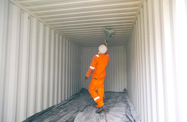 Wide view of a person in an orange protective suit spraying insulation coating onto the ceiling of a shipping container, with protective sheeting covering the floor. 
