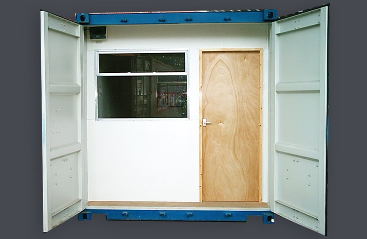 Modified shipping container with double cargo doors open, revealing an internal plywood partition wall and fitted hardwood personnel door with a window installed beside it. 