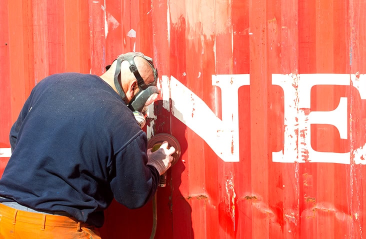 Worker wearing a respirator sanding the exterior surface of a red shipping container, removing paint and preparing the metal for refurbishment