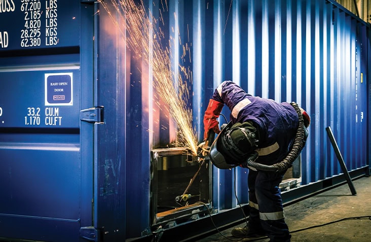 Engineer welding a cut-out section into the side of a blue shipping container, creating sparks during container modification work. 