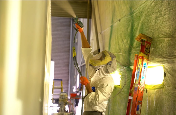 A person wearing protective coveralls, gloves, and a respirator is spraying paint onto a vertical surface inside a paint booth. A red ladder and bright work lights are visible in the background, along with green protective sheets covering the walls. 