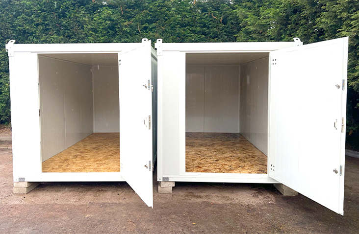 Two white insulated flat pack storage units placed side by side outdoors, both with open doors showing plywood flooring and insulated interior walls, designed for secure storage.