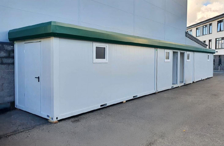 Interior of a modular building with white walls, grey flooring, ceiling lights, and large windows allowing natural light to fill the space.