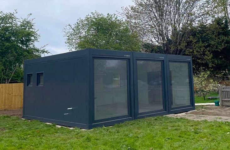 Dark grey modular nursery building with large floor-to-ceiling windows and small side windows, installed on a grassy site surrounded by trees and fencing.