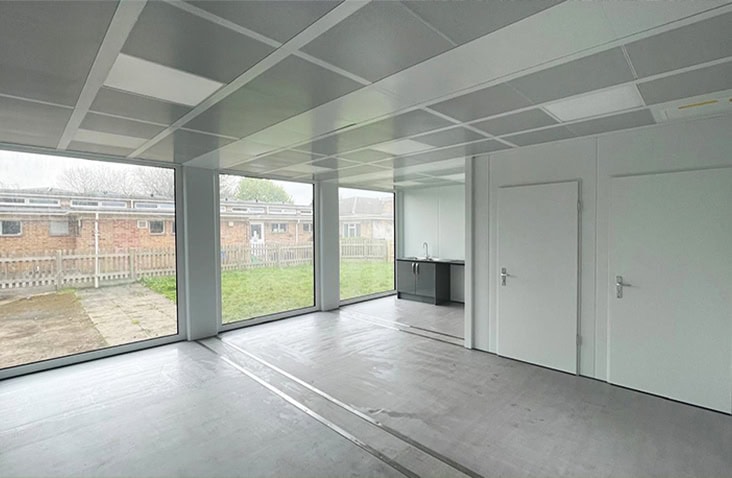 Interior of a modern modular classroom with large floor-to-ceiling windows, white walls, grey flooring, and a small kitchenette area near the back.