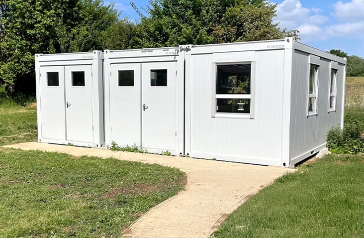 White modular classroom building made from three connected units with multiple doors and windows, situated on a school field with a paved path leading to the entrance.