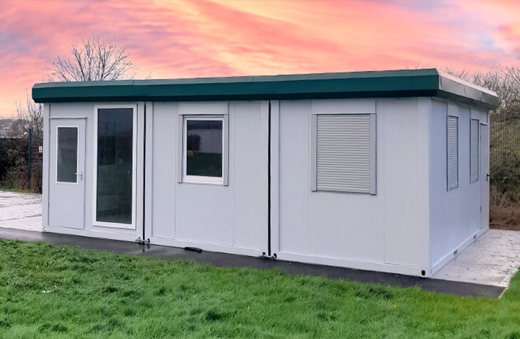 White modular building with green roof trim, large windows fitted with shutters, and a glass entrance door, installed on a concrete base with grass in the foreground under a pink sunset sky.