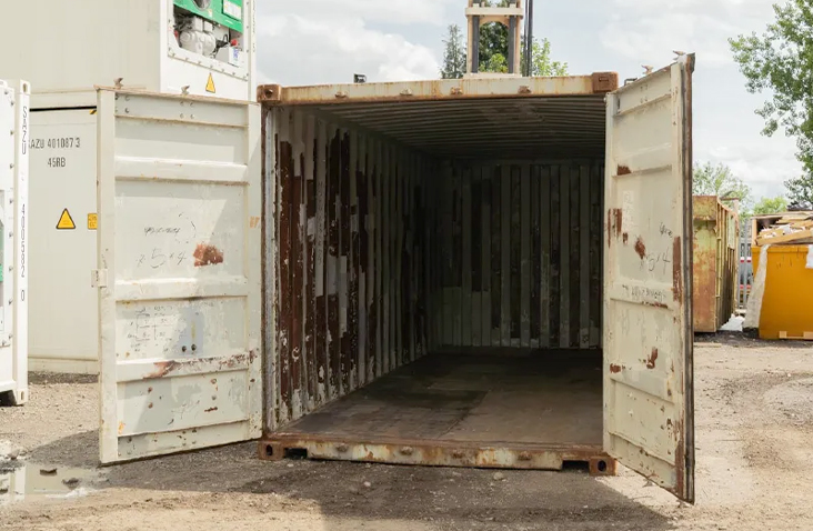 Used 20ft shipping container in light grey with both cargo doors open, showing a worn but dry steel interior with visible rust and paint wear, suitable for low-cost storage on farms, construction sites, or industrial yards. 
