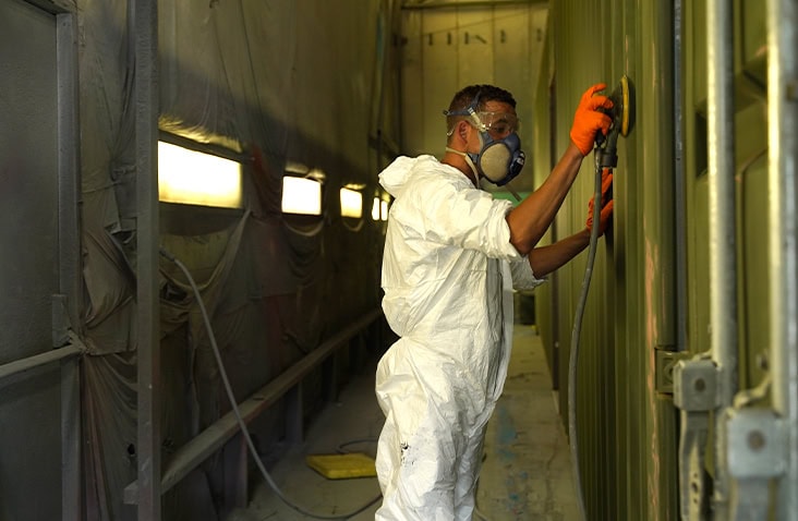 Worker wearing white overalls, gloves, and a respirator mask preparing a green shipping container for repainting inside a paint booth. The worker sands the container’s exterior while standing next to bright overhead panel lighting. 