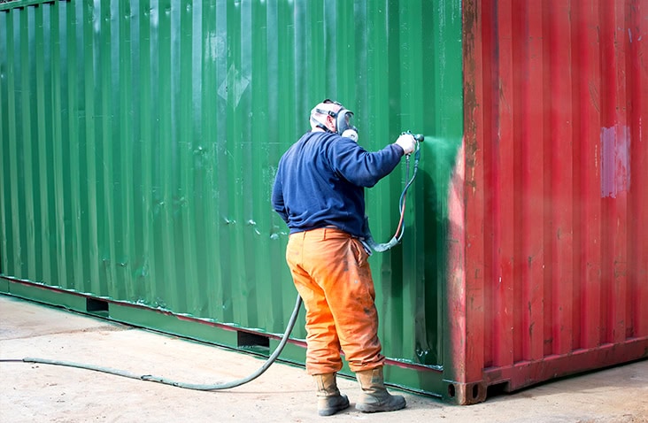 Worker wearing a respirator and protective clothing spray-painting the side of a shipping container, applying green paint over a previously red surface