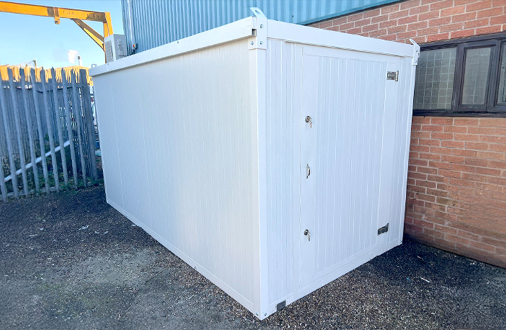 White insulated flat pack storage unit with single door, installed beside a brick building and metal fencing on an industrial site, designed for secure and temperature-controlled storage.