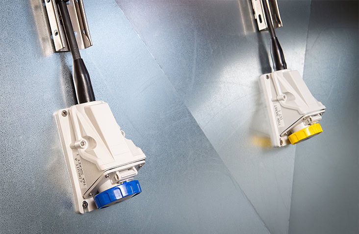 Industrial IEC 60309 panel-mounted power sockets in blue and yellow fitted to a metal-lined shipping container wall. 