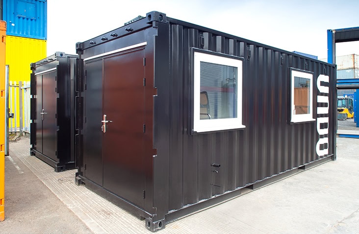 Black converted container with two windows and double personnel doors, located in a container depot with other coloured containers nearby. 