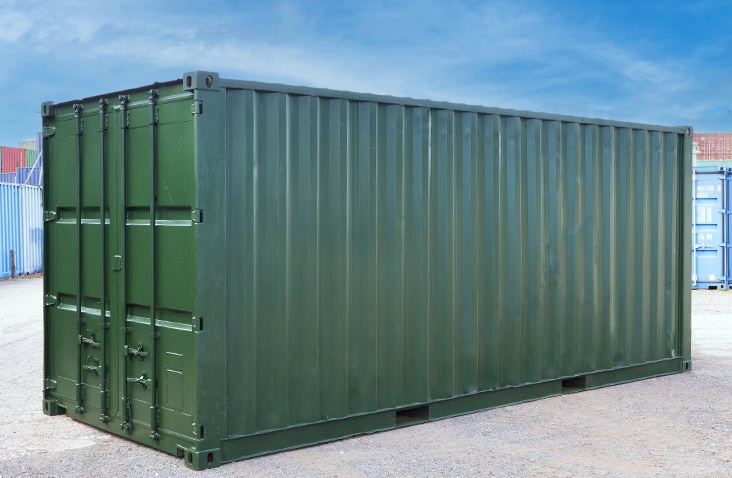 Green steel shipping container with corrugated sides and closed double doors, placed on a gravel surface outdoors with other containers visible in the background under a clear blue sky. 