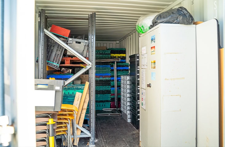 Interior of a 20ft shipping container used for storage, filled with shelving, plastic crates, stacked trays, and assorted equipment. 