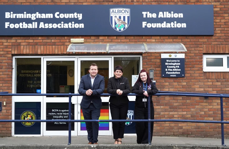 Three people standing outside the Birmingham County Football Association and Albion Foundation building entrance, beneath exterior signage