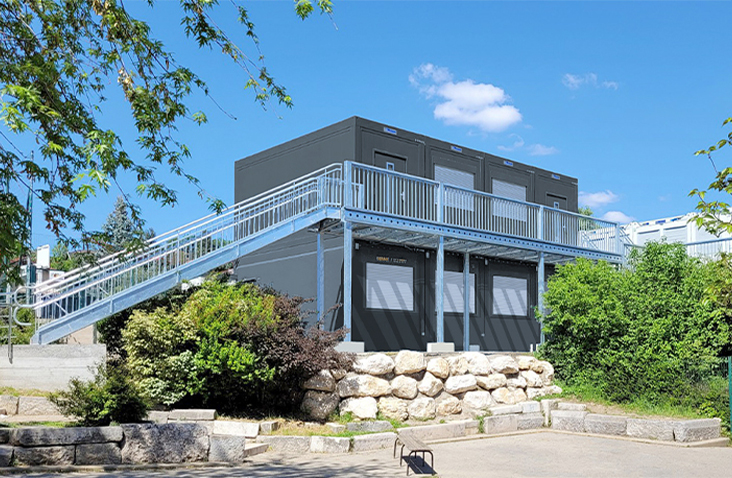Two-storey modular building constructed from stacked units with external metal stair access and railings, surrounded by trees and landscaping under a blue sky.